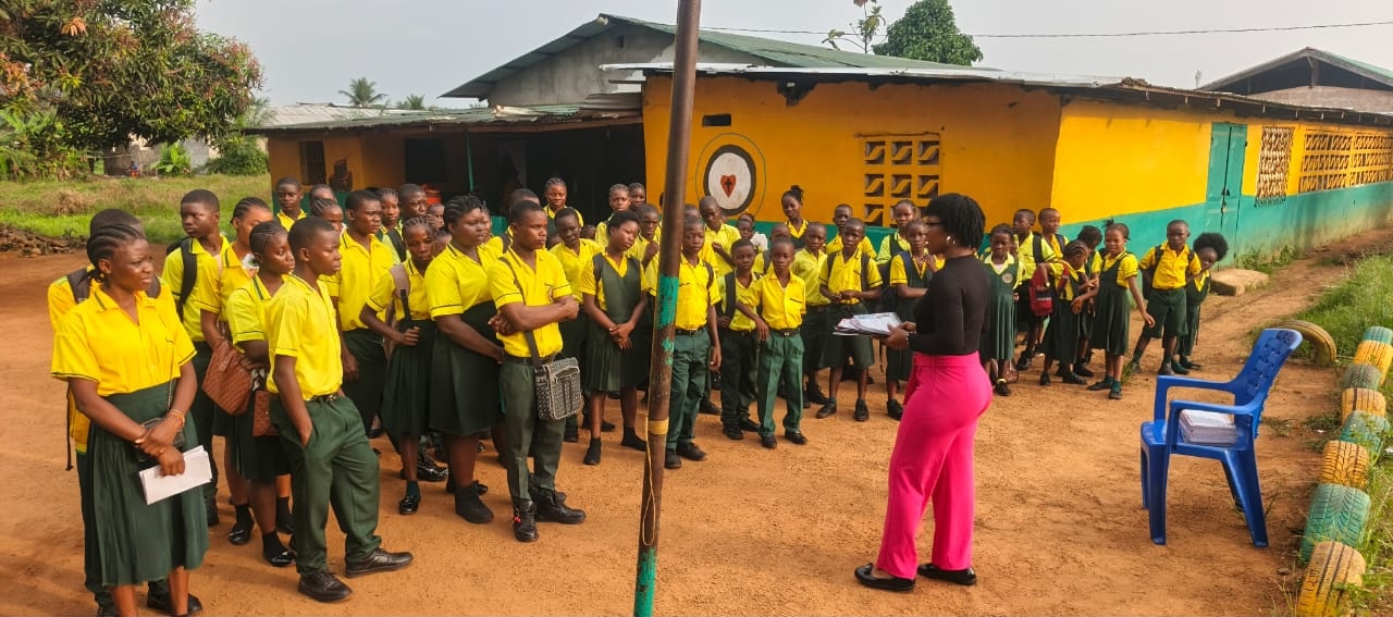 Teacher in pink addressing students in yellow and green uniforms during an outdoor school assembly